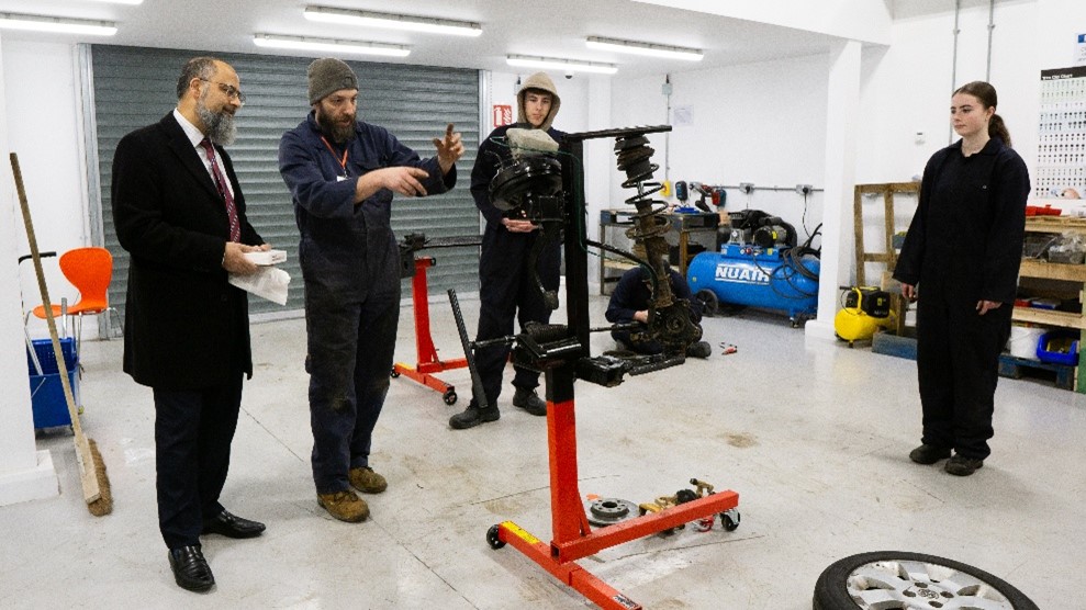 A group of people in a workshop, one man in overall explains something, gesturing to a car part, with two students and another man watching.