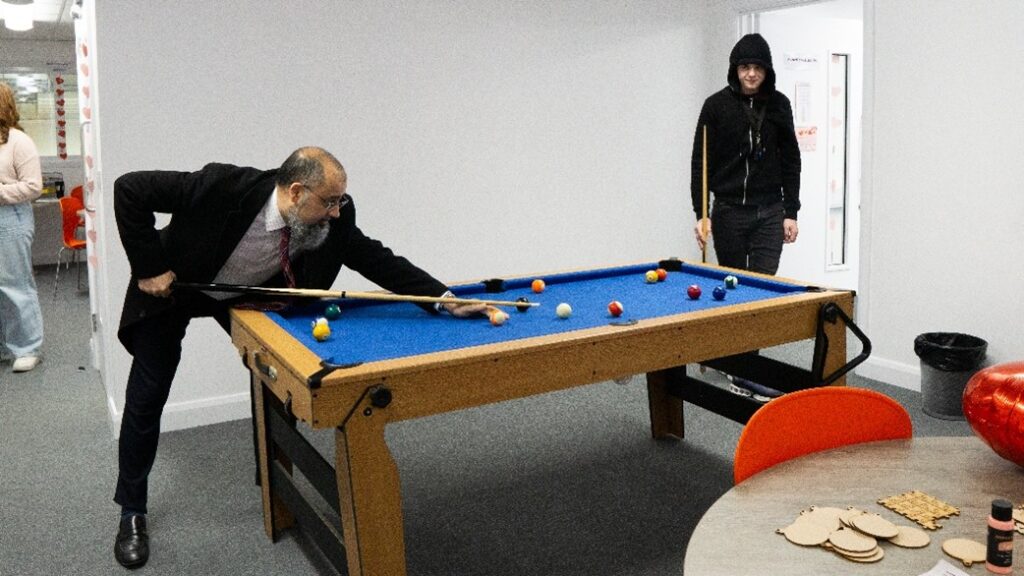 A blue pool table, two people are playing with one person leant over the table, taking a shot while the other person watches them