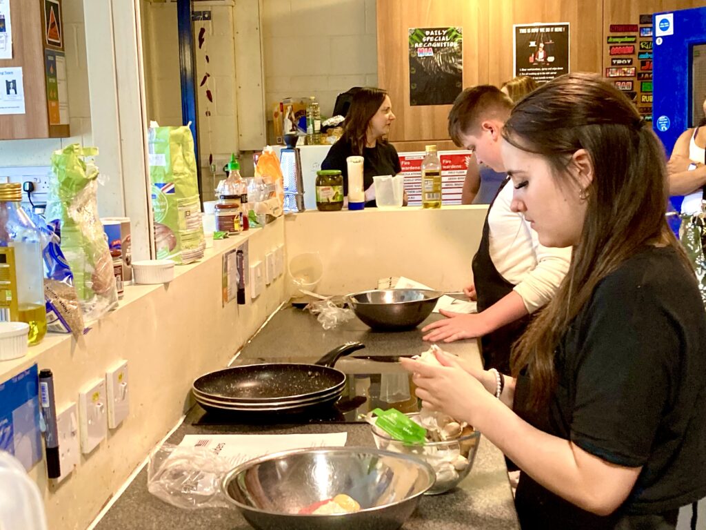 A group of people cooking in a communal kitchen setting. In the foreground, a woman is peeling mushrooms over a metal bowl, while others are busy with food preparation along a counter lined with ingredients and utensils. A mirror reflects the activity, and posters and kitchen supplies are visible in the background.