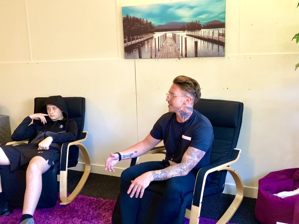 Two people sit in a relaxed setting on black cushioned chairs. One person, a young man in a black hoodie and shorts, appears quiet and thoughtful. The other person, an adult with visible tattoos and glasses, is speaking and gesturing with one hand. A scenic photograph of a wooden dock over water with mountains in the background hangs on the wall behind them.