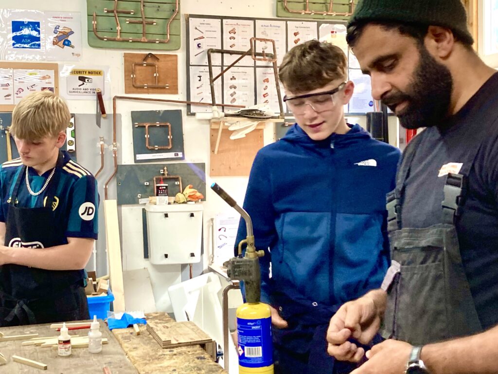 Three individuals in a workshop setting engaged in a hands-on activity. One adult, wearing a beanie and overalls, appears to be instructing or demonstrating something to two teenage boys. One boy is wearing a blue sports jersey and apron, while the other, in safety goggles and a blue hoodie, looks on attentively. Various tools, wooden pieces, and copper piping displays are visible in the background.