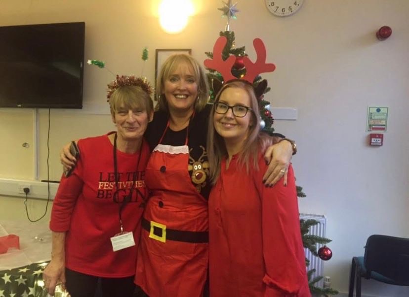 The image shows three women posing for the picture, it looks like it is Christmas as there is a tree and presents in the background, they are all dressed in red and all have blonde hair.