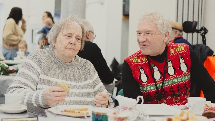 The image shows two people, a woman sat on the left and a man sat on the right, they are sat a table that is dressed with plates, food and teacups. The woman is eating a sandwich, and wears a white and grey striped jumper. The man smiles at her whilst wearing a Christmas jumper that is red with penguins and reindeer on. There are people chatting in the background.