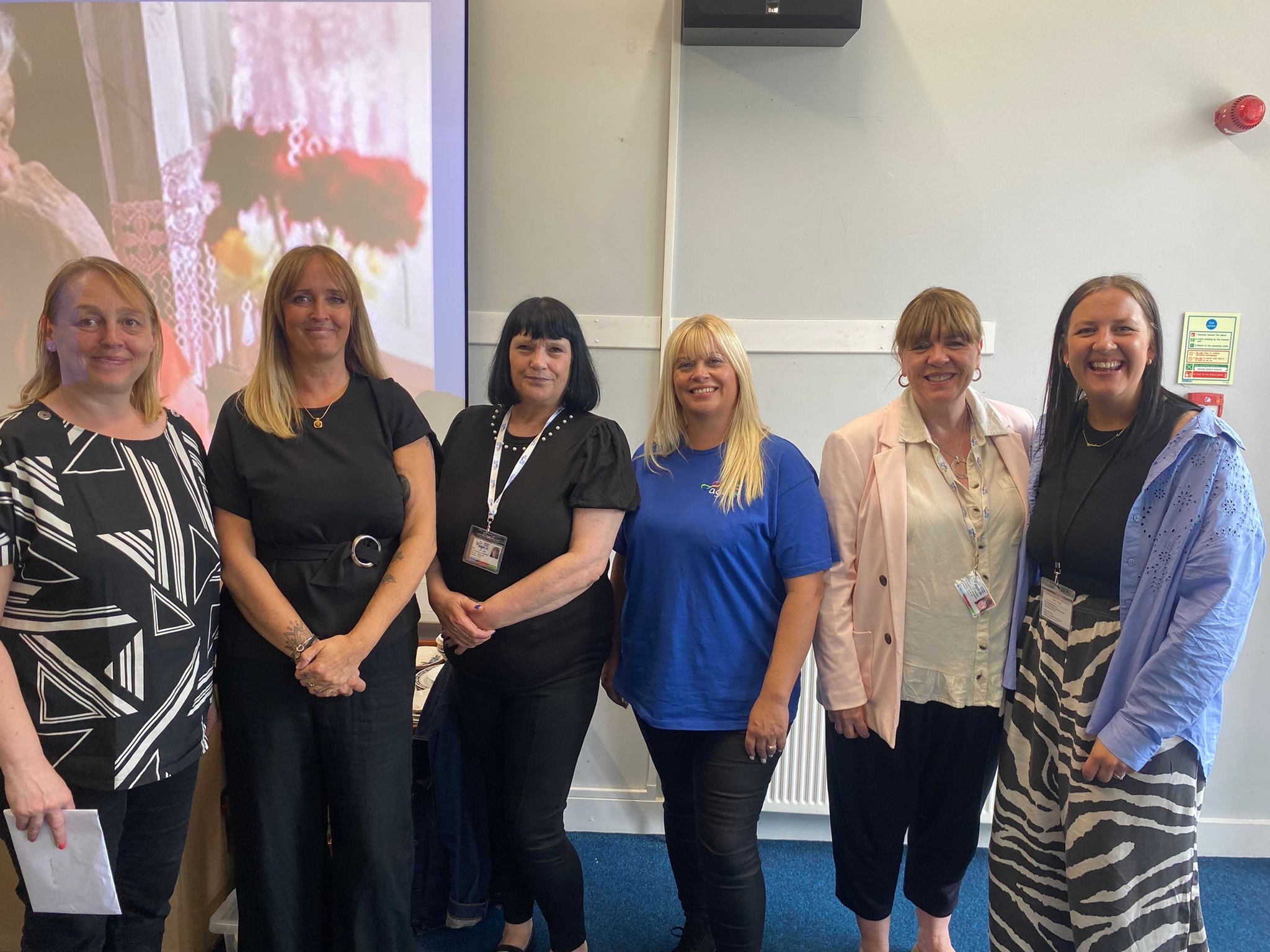 The image shows six women, stood in a line all posing for the picture. The background looks like a classroom with a projection on the wall in the lefthand half of the picture. The women range from 30s to middle aged and they are all smiling at the camera.