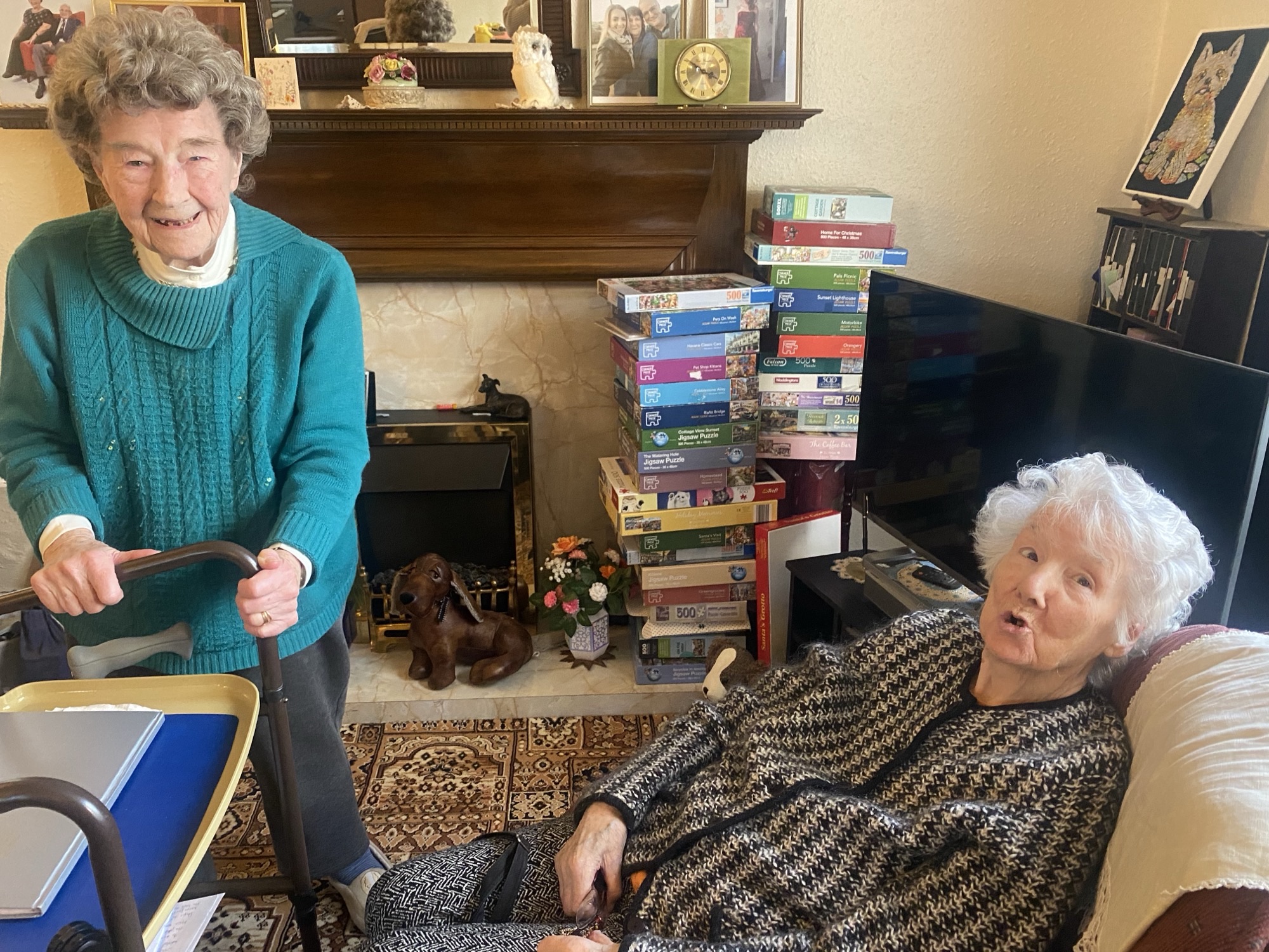 The image shows two women in a living rooms, there is a fireplace and a stack of video tapes in the background. The room is decorated in beiges and browns. One of the women is on the right, sat in a comfy chair and appears to be in the middle of saying something. The other woman is sat behind a elevated dinner tray and is laughing, she is wearing a teal jumper.