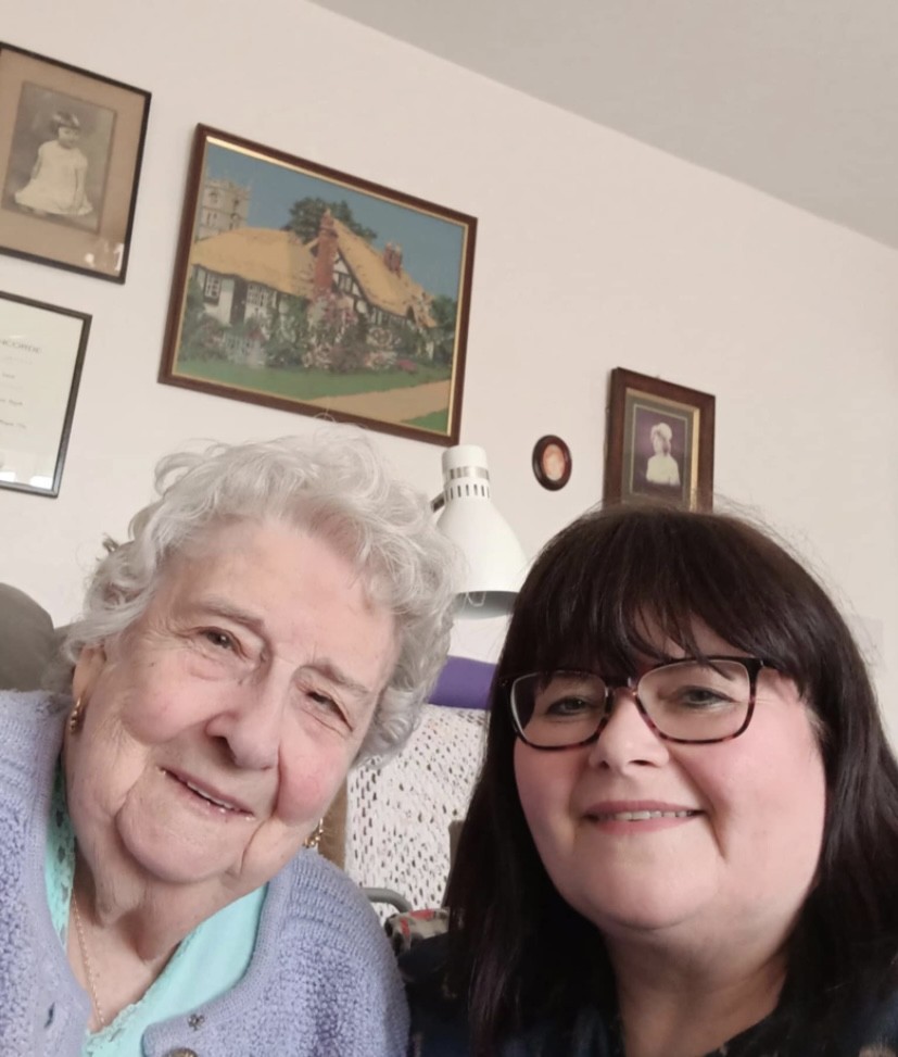 The image shows two women smiling at the camera, it looks like a living room setting with pictures on the wall. The woman on the left is elderly, with white hair and wears a purple cardigan, the woman on the right has dark hair, with a fringe and dark rimmed glasses.