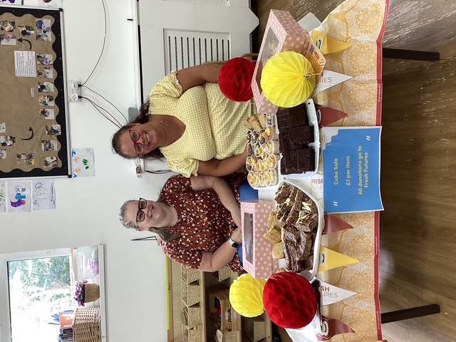 Two smiling women sit behind a cake sale table decorated with red and yellow paper ornaments, offering a variety of cakes and bakes to raise funds for Fresh Futures.