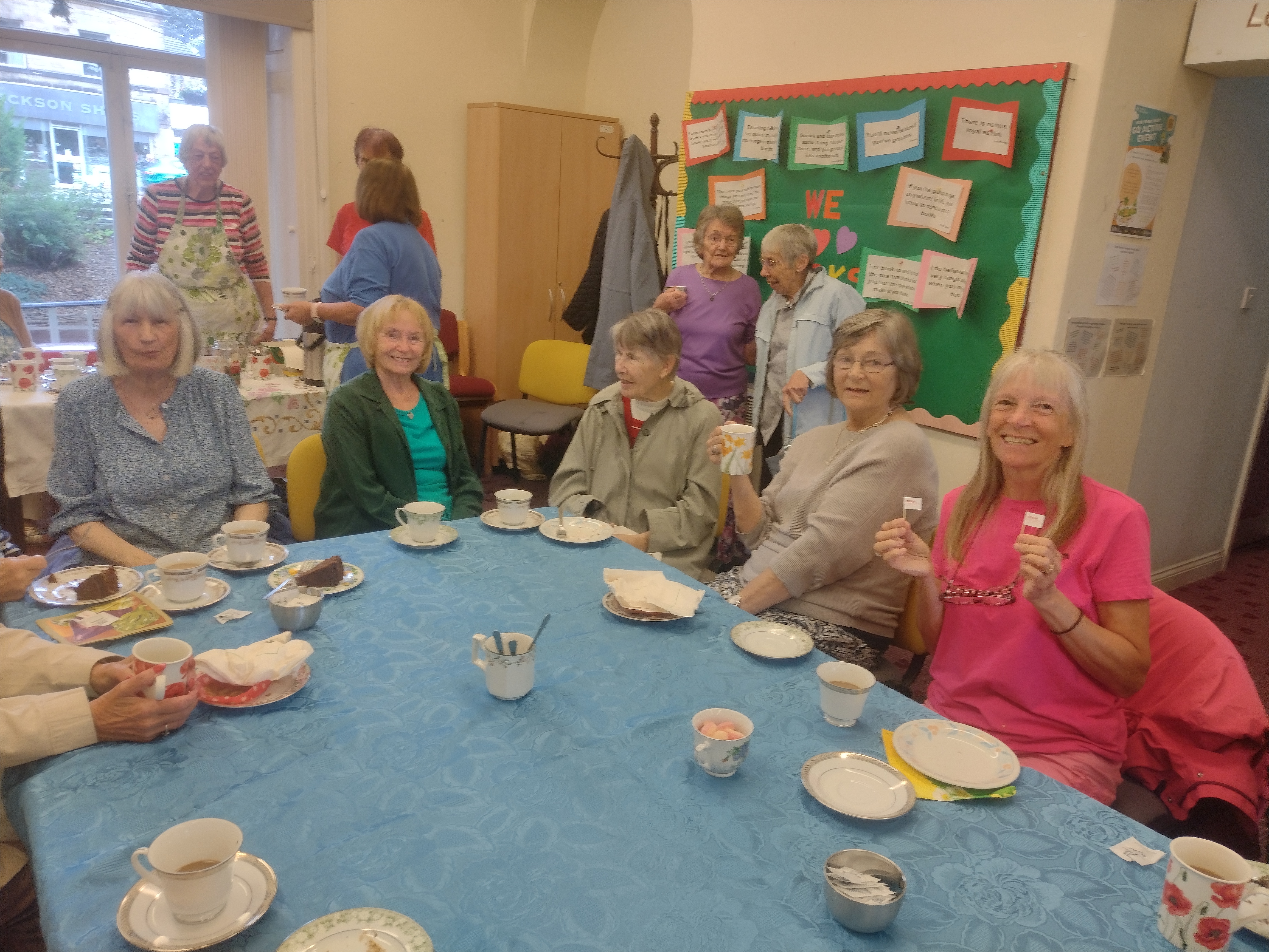 A group of older women sit around a blue tablecloth enjoying tea, cake, and conversation at a community coffee morning, with more people chatting in the background.