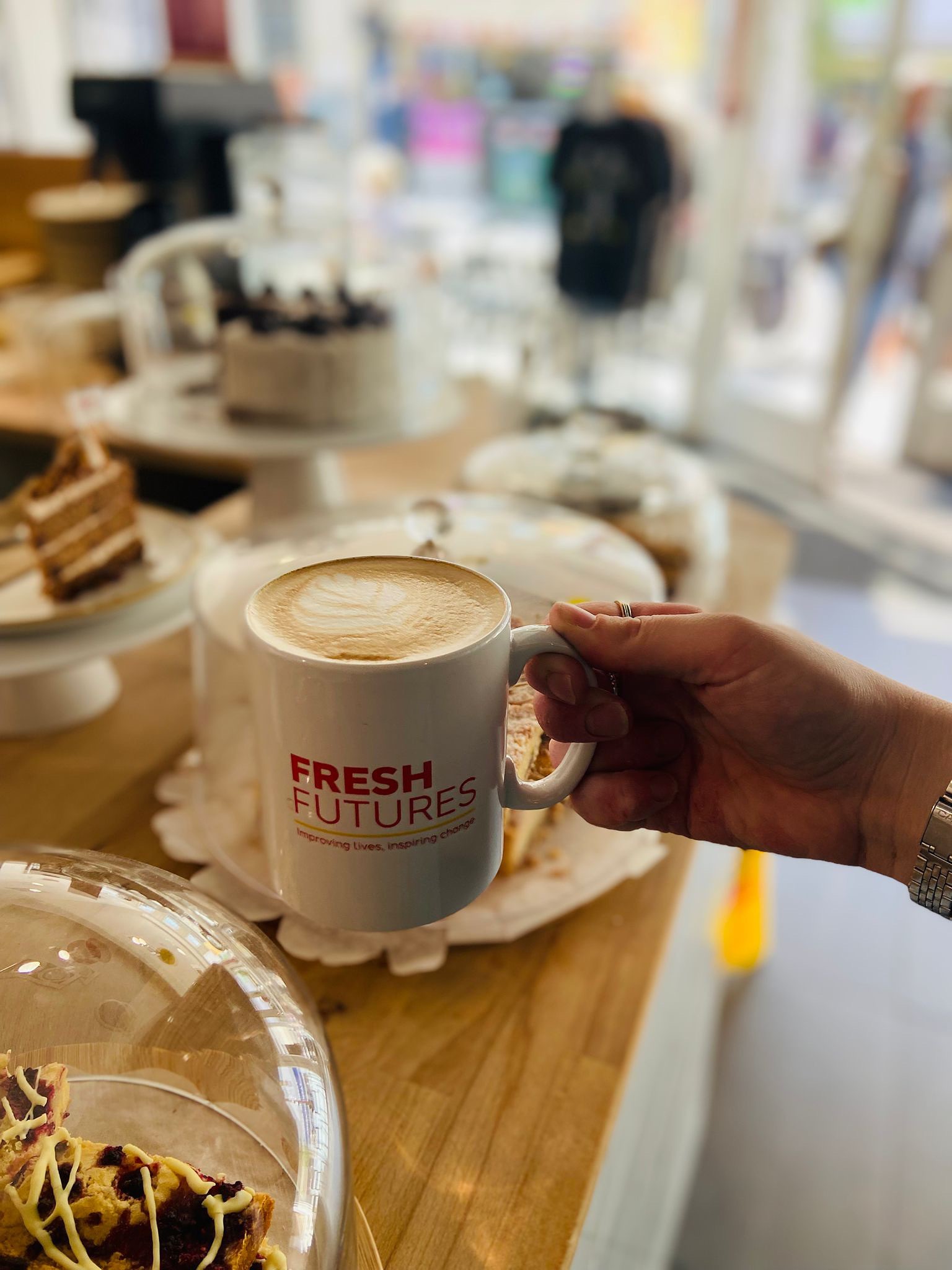  A close-up of a hand holding a cappuccino in a Fresh Futures mug, with cakes and slices displayed under glass covers in the background.