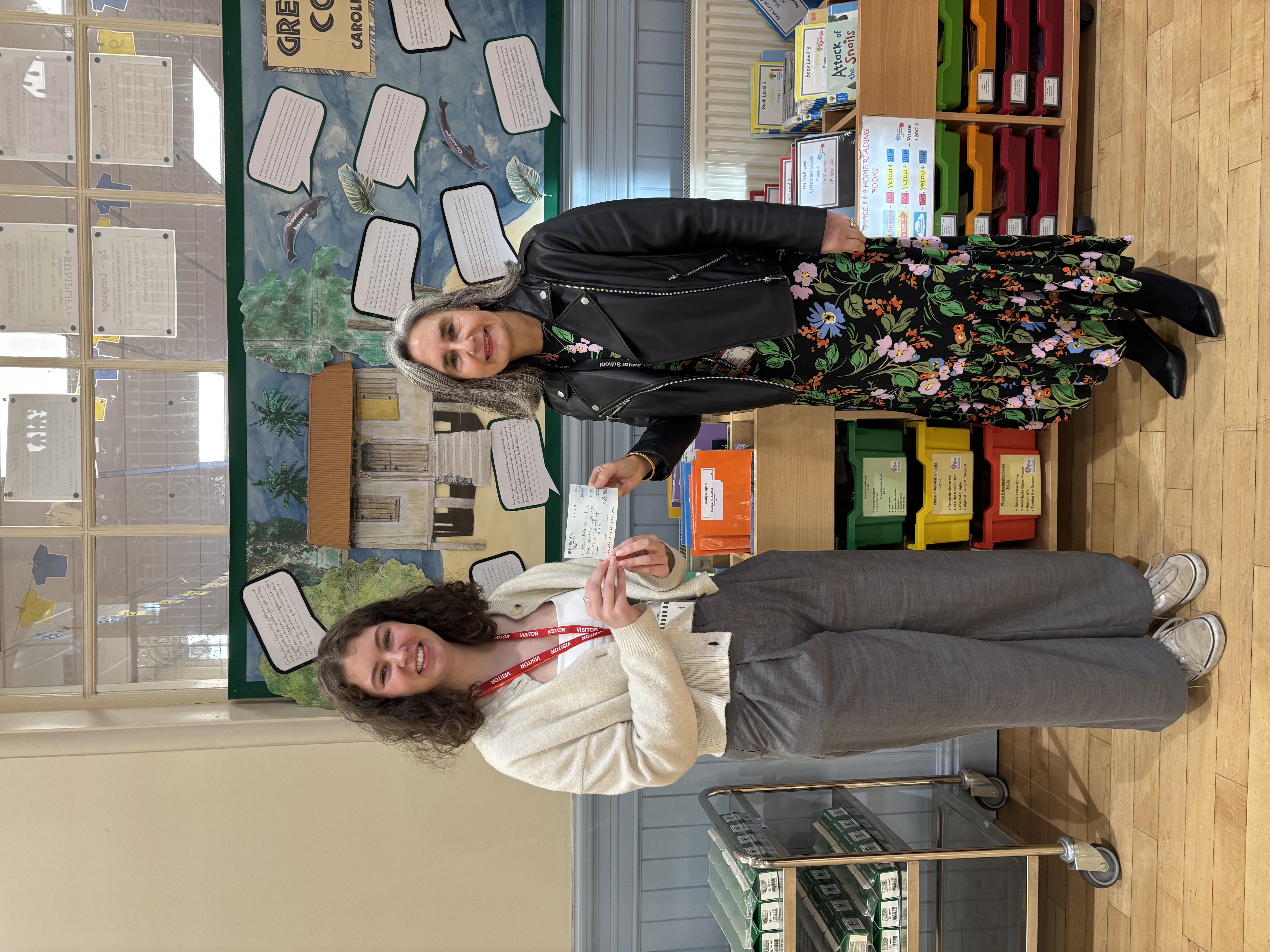Two women stand smiling indoors as one hands the other a cheque. They are in front of a colourful classroom display featuring a painted tropical house scene with written notes around it. One woman wears a white cardigan and grey trousers, and the other wears a black leather jacket over a floral dress. They appear to be marking a donation or presentation moment in a cheerful school setting.