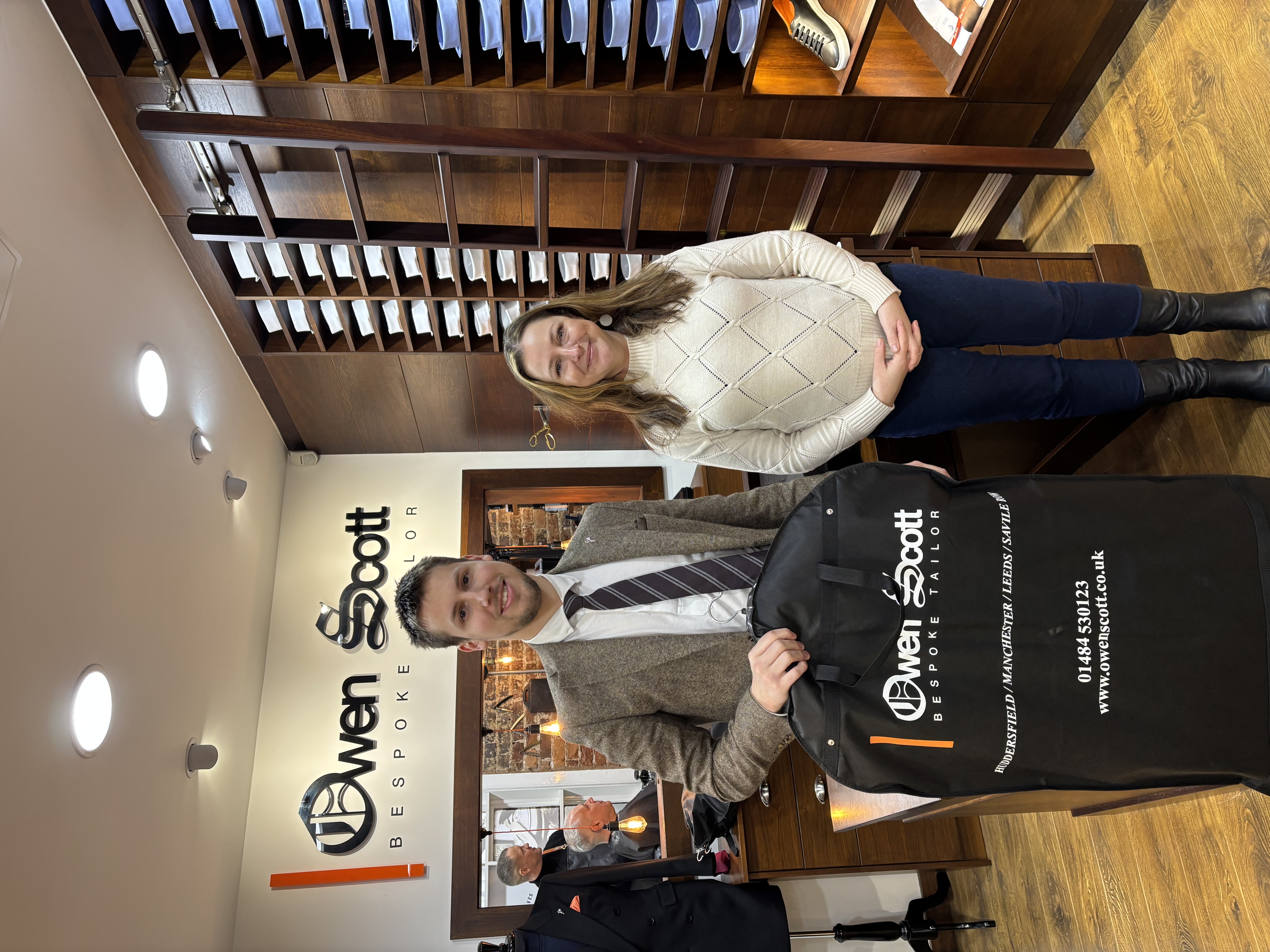 Two people stand inside a bespoke tailor shop with warm wooden interiors. A man in a tweed jacket and tie holds a black garment bag branded “Owen Scott Bespoke Tailor,” while a woman in a cream knit sweater and dark jeans stands beside him, smiling. Behind them are shelves neatly stacked with folded shirts and a wall sign reading “Owen Scott Bespoke Tailor,” with mirrors and tailoring displays visible in the background.Two people stand inside a bespoke tailor shop with warm wooden interiors. A man in a tweed jacket and tie holds a black garment bag branded “Owen Scott Bespoke Tailor,” while a woman in a cream knit sweater and dark jeans stands beside him, smiling. Behind them are shelves neatly stacked with folded shirts and a wall sign reading “Owen Scott Bespoke Tailor,” with mirrors and tailoring displays visible in the background.